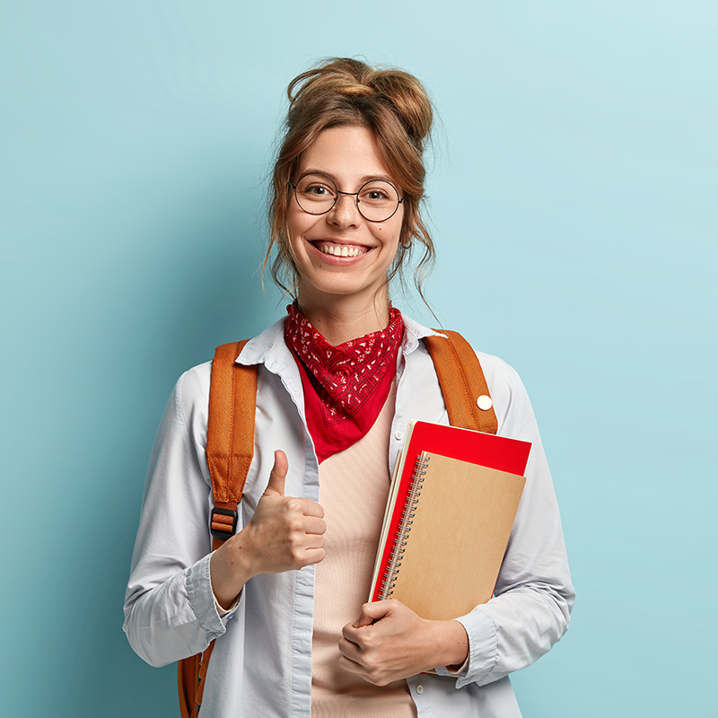 Glad student gives approval, keeps thumb up, holds notebook and diary, has happy smile, says okay, wears optical spectacles, casual outfit, studies at college, isolated over blue background.